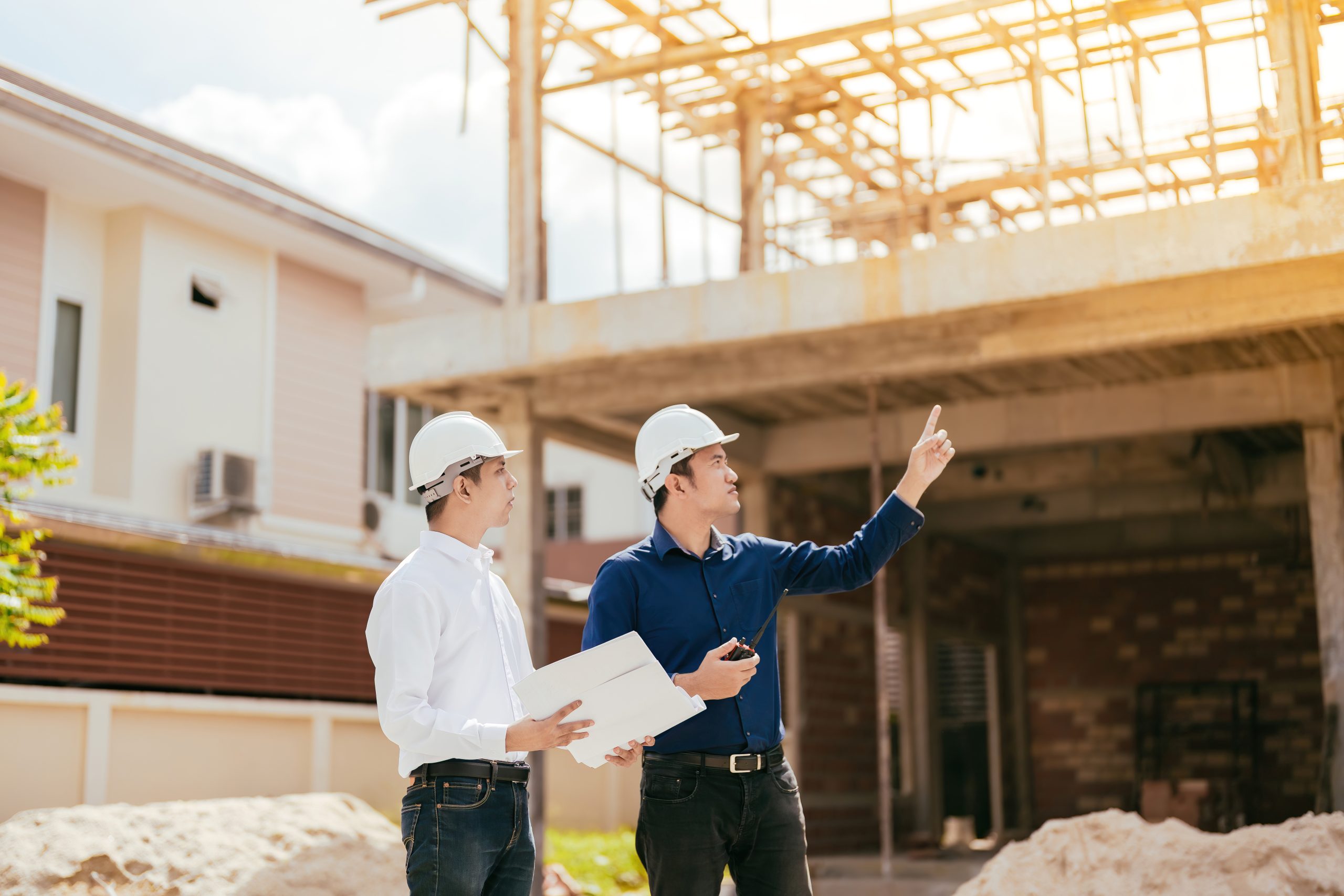 Two male engineers, Asians, are the technicians. Pointing to a newly built housing project related to the architectural industry, holding a blueprint, radio, wearing a hard hat and uniform.